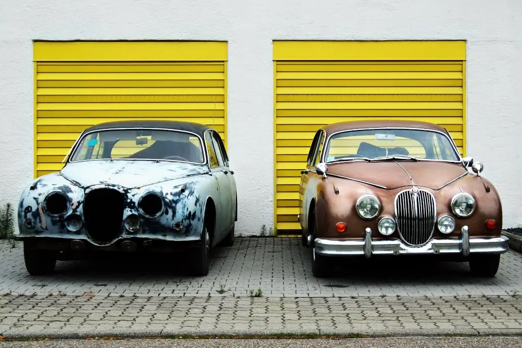 Two vintage Jaguars parked in front of a sunny-yellow garage.