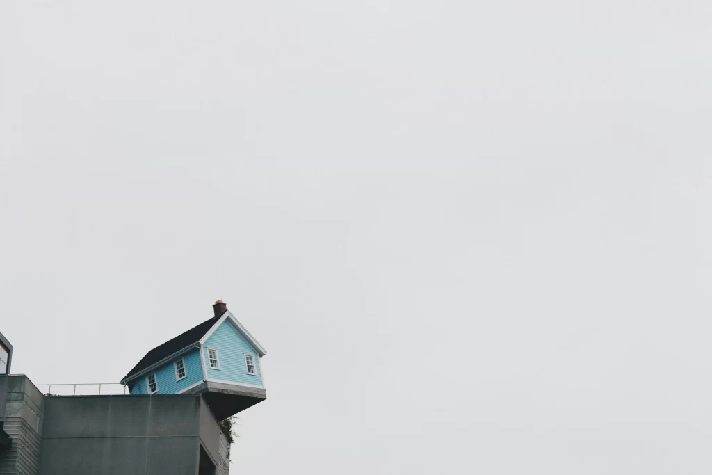 A small blue house appears to precariously balance on the edge of a modern building under a cloudy sky.