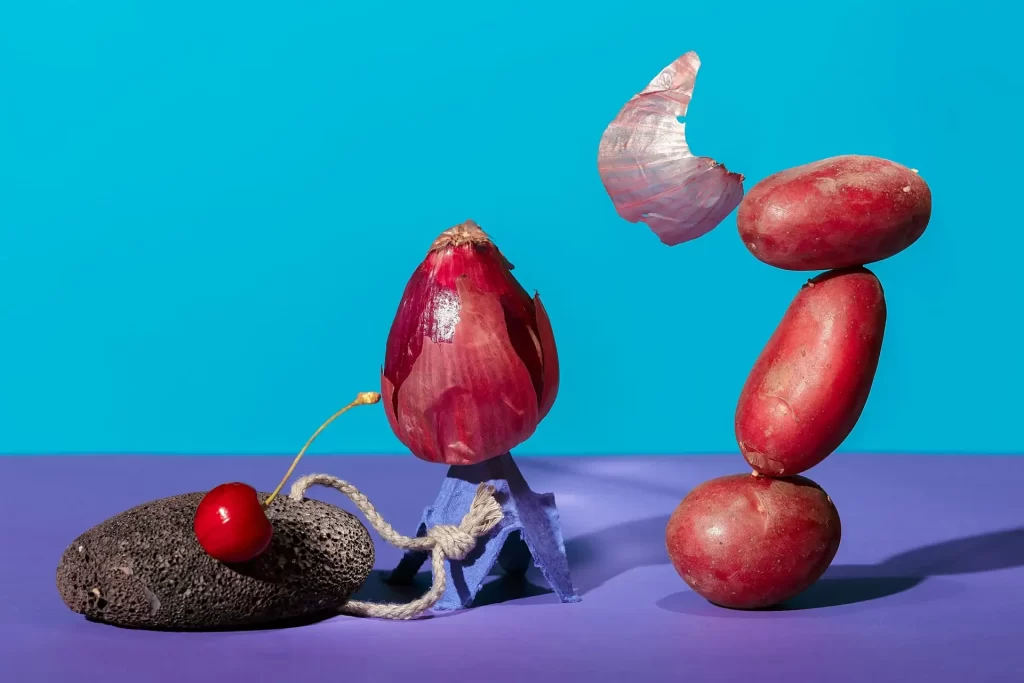 An arrangement of a cherry, avocado, onion, and three red potatoes in a precarious balance against a bright blue and purple background.